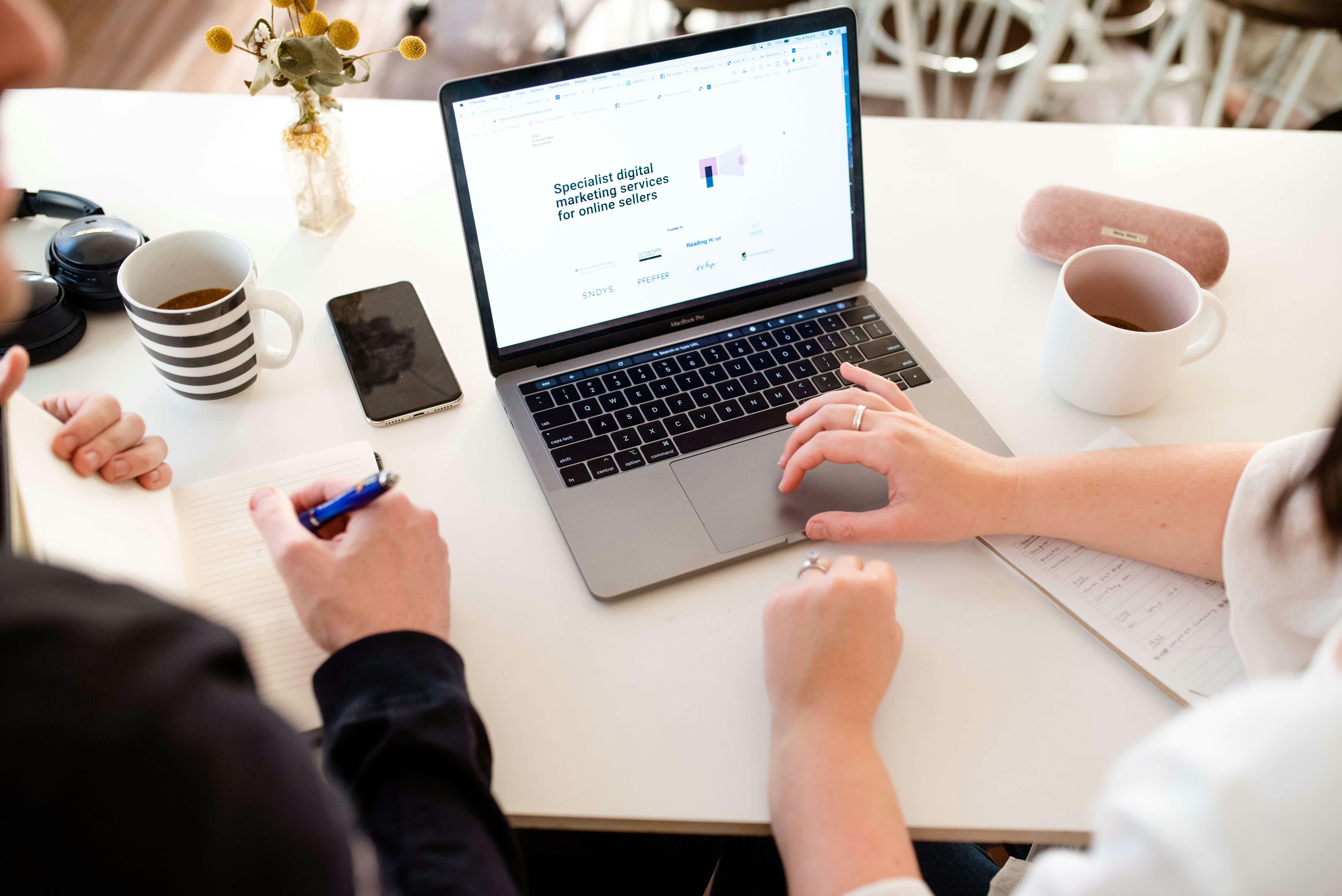 A person's hand typing on a laptop keyboard with a coffee cup beside it displaying the text "Chrome, Bookmarks, People, Window, Help, 100%, Down, X, Options, Ad Library, The Connected Narrative, Careers, Welcome,